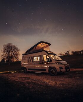 Carado motorhome with the pop-up roof raised at night on an open parking spot under a starry sky