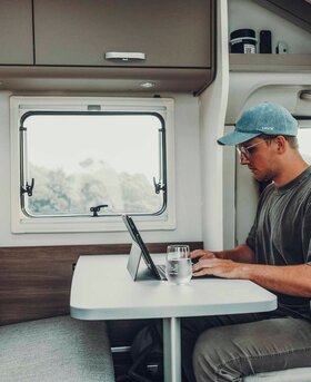 A man working in a Carado motorhome at the dinette with a table, tablet and window inside.
