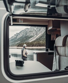 View from a Carado motorhome of a man by the lake with a mountain backdrop; interior with table and kitchen