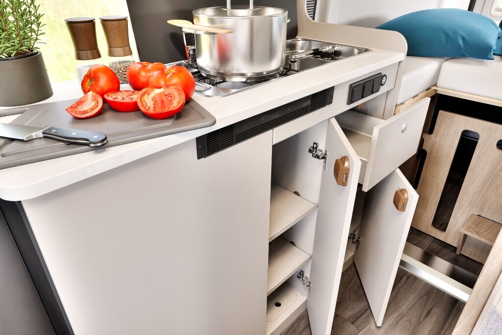 Küchenzeile im Carado Wohnmobil mit Gaskocher, Spüle, Schneidebrett mit Tomaten und geöffneten Staufächern