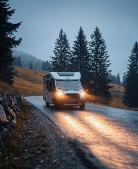 Carado camper met de verlichting aan, rijdend over een natte landweg in de schemering, omringd door sparren en heuvels