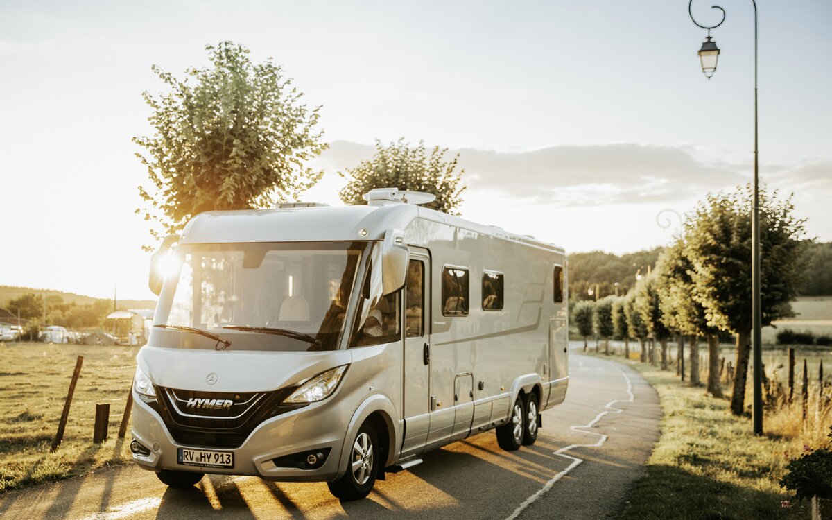 Doppelachser HYMER B-Klasse MasterLine I fährt entlang einer von Bäumen gesäumten Straße in warmer Lichtstimmung