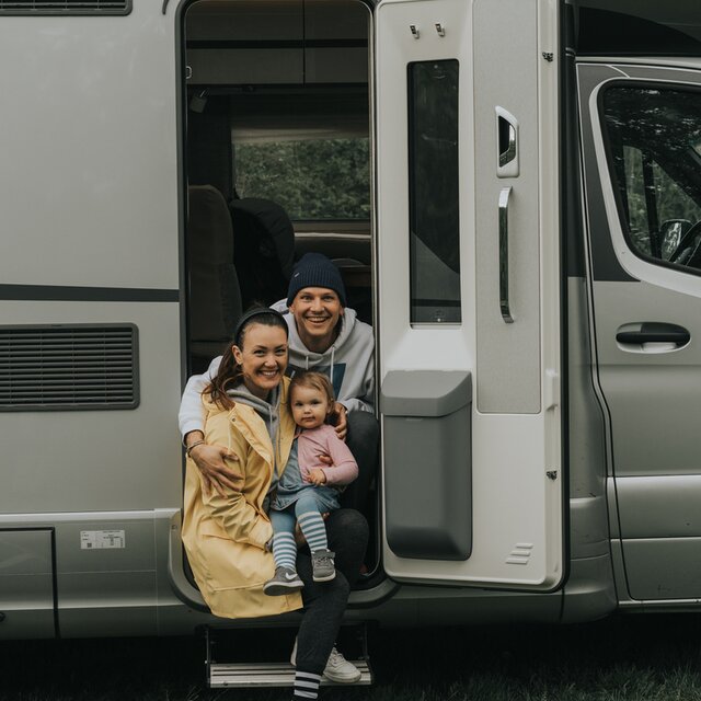 Junges Paar sitzt mit Kleinkind in der geöffneten Aufbautüre eines Reisemobils