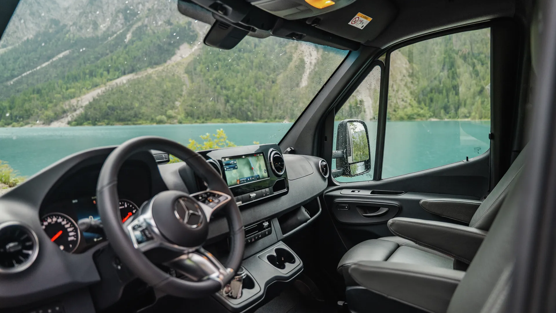 Fahrerhaus des Hymer B‑ML T mit komfortablen Pilotensitzen, modernem Cockpit und beeindruckendem Blick auf einen Bergsee inmitten alpiner Natur