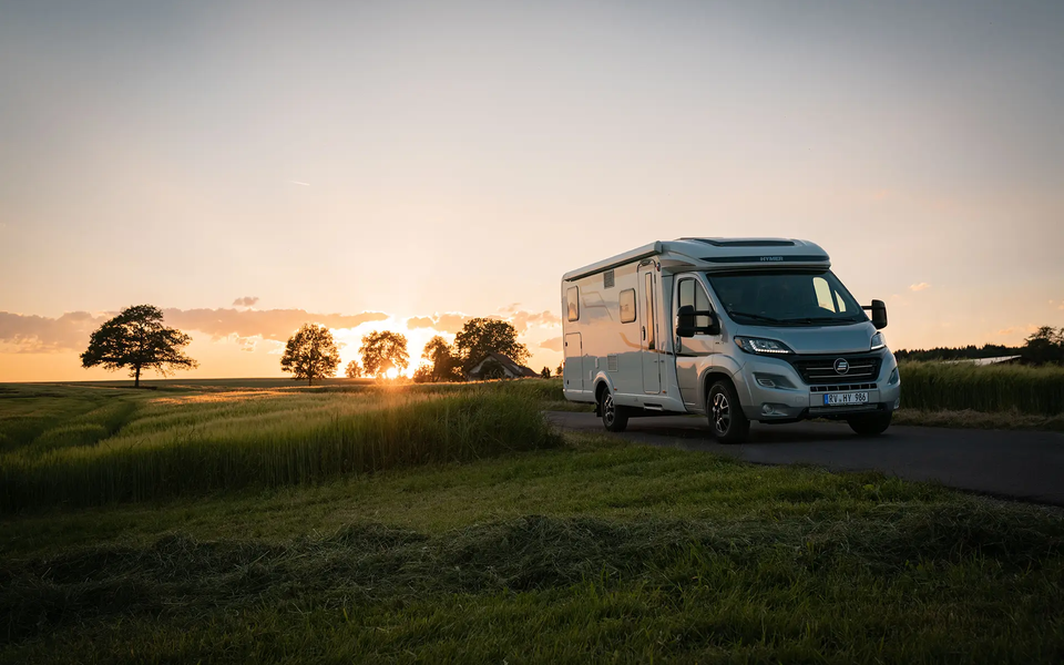 HYMER Exsis-t auf schmaler Straße inmitten grüner Wiesen und Kornfelder bei Sonnenuntergang