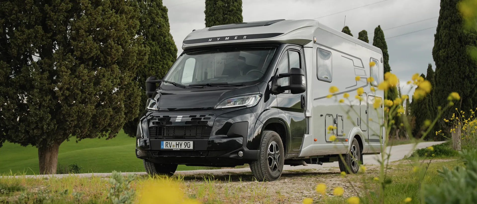 Der Hymer Exsis‑t mit schwarzem Fahrerhaus und silbernem Aufbau steht auf einem Feldweg in ländlicher Landschaft mit Zypressen, Blumen im Vordergrund und bewölktem Himmel.