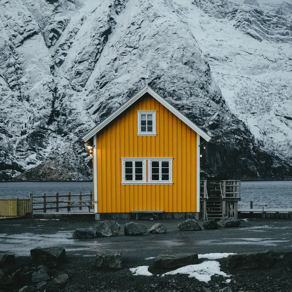 Lite gult hus ved fjorden, et stort snødekt fjell i bakgrunnen