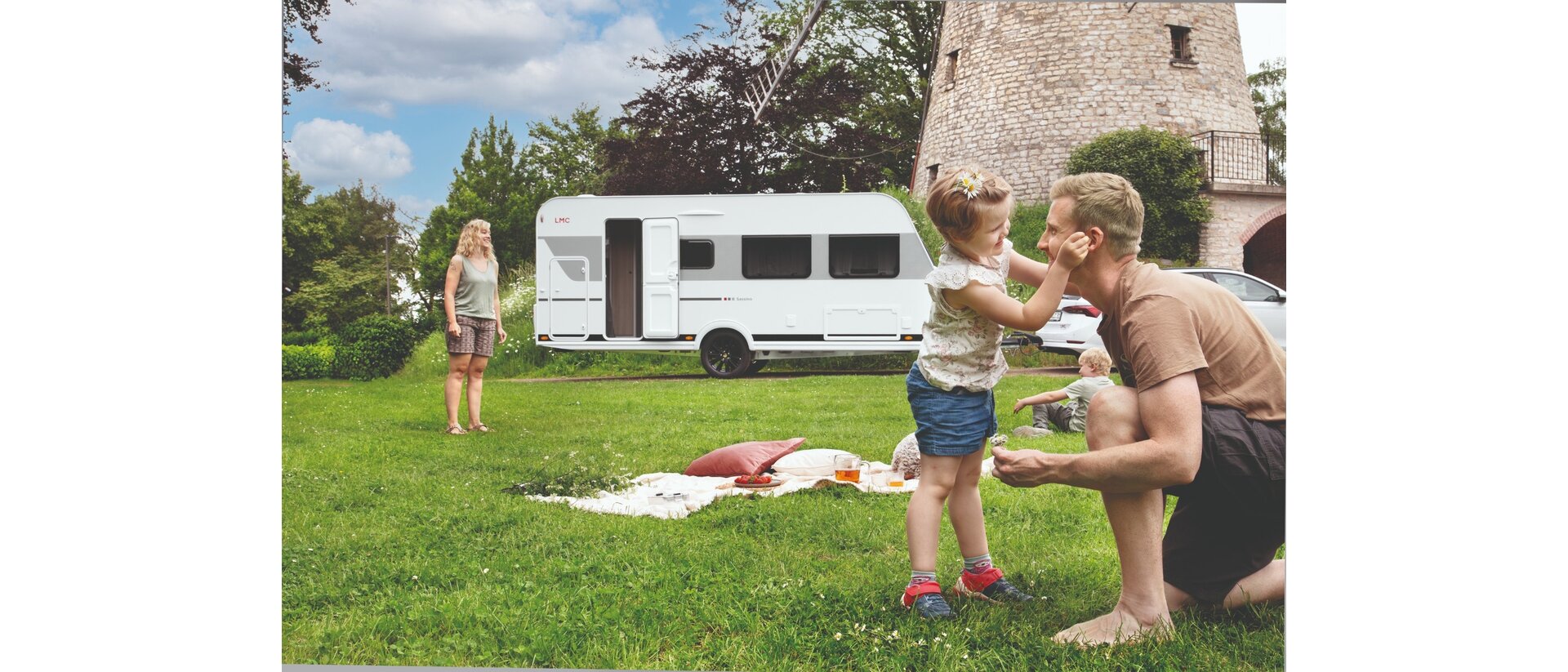 De foto toont een jong gezin dat geniet van een picknick op een groene weide naast de LMC Sassino-caravan en een molen.